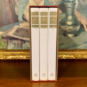 May include: A three-volume set of "The Divine Comedy" by Dante Alighieri. The books are bound in white with gold lettering and are arranged in a red slipcase. The titles of the books are "Paradise", "Purgatory", and "Hell".