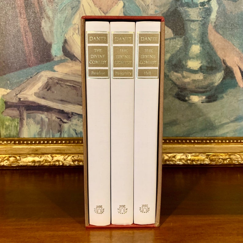 May include: A three-volume set of "The Divine Comedy" by Dante Alighieri. The books are bound in white with gold lettering and are arranged in a red slipcase. The titles of the books are "Paradise", "Purgatory", and "Hell".