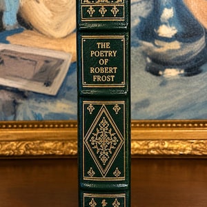 Vintage Leather-bound edition of The Poetry of Robert Frost in dark green with ornate gold gilt decoration on the cover, standing upright on a wooden surface in front of a framed painting.