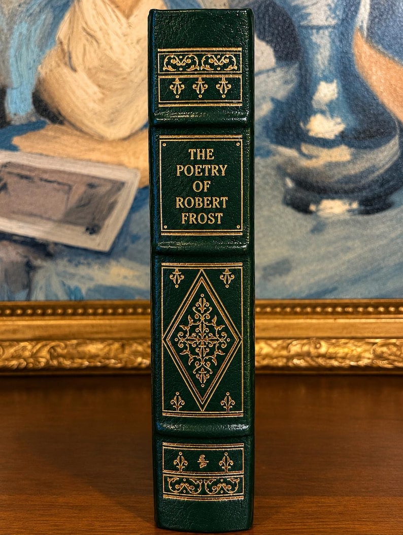 Vintage Leather-bound edition of The Poetry of Robert Frost in dark green with ornate gold gilt decoration on the cover, standing upright on a wooden surface in front of a framed painting.