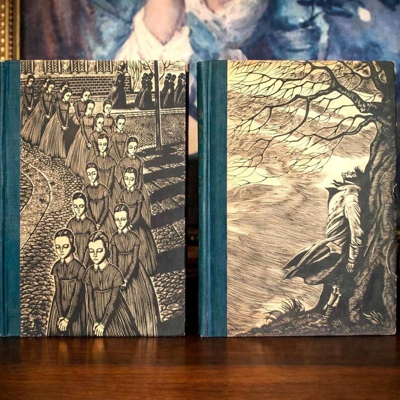 Peut inclure: Deux livres reli&eacute;s avec des dos bleu sarcelle fonc&eacute; et des couvertures cr&egrave;me orn&eacute;es d'illustrations &agrave; l'encre noire. Le livre de gauche montre une procession de filles en robe sur un chemin pav&eacute;. Celui de droite montre une personne adoss&eacute;e &agrave; un arbre.