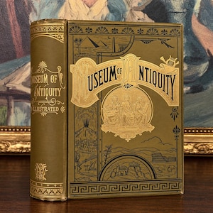 May include: A hardcover book titled Museum of Antiquity Illustrated with a dark olive green cover and gold foil stamping. The front cover features an illustration of three figures, pyramids, and ancient architecture with decorative borders.