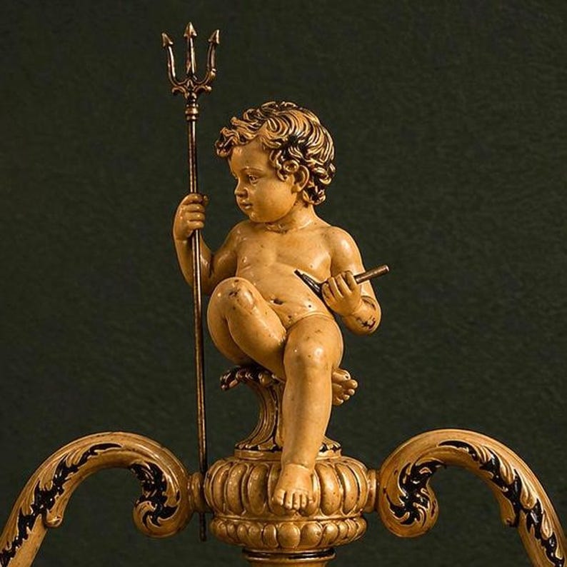 K&ouml;nnte beinhalten: Eine beige Puttenfigur mit lockigem Haar, die auf einem dekorativen Sockel sitzt. Die Figur h&auml;lt einen hohen, d&uuml;nnen Dreizack in der rechten Hand und ein kleines, dunkles, spitzes Werkzeug in der linken Hand. Der Sockel zeigt geschwungene Ornamente.