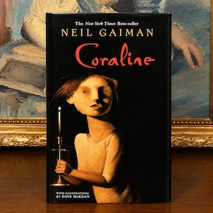 May include: A hardcover book titled Coraline by Neil Gaiman with a black dust jacket. The cover features an illustration of a pale figure holding a lit candle against a dark background. The text reads The New York Times Best-seller and With Illustrations by Dave McKean.