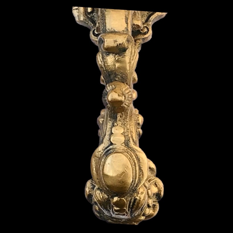 May include: Ornate brass-colored decorative object with a detailed, symmetrical design. The object features floral and oval embellishments, with a textured surface. The piece has a vintage aesthetic, suitable for home decor or restoration projects.