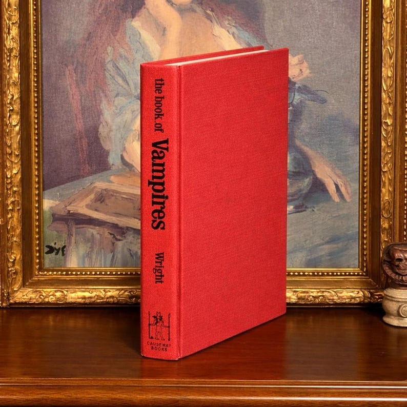 May include: A hardcover book titled "the book of Vampires" by Wright. The book has a bright red cover with white lettering and a small illustration. The book is standing upright on a wooden surface.