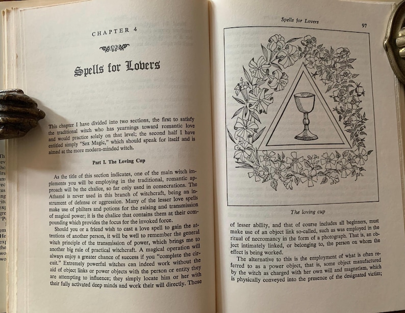 Puede incluir: Un libro abierto con el t&iacute;tulo "Spells for Lovers" en la p&aacute;gina izquierda. La p&aacute;gina derecha presenta una ilustraci&oacute;n floral que rodea un c&aacute;liz dentro de un tri&aacute;ngulo. El texto detalla el enfoque del cap&iacute;tulo en hechizos de amor.