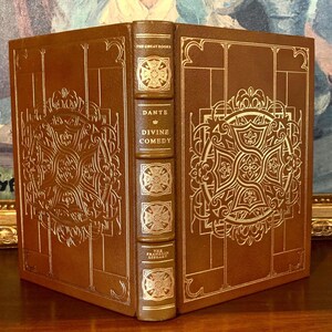 Leather-bound edition of Dante's 'Divine Comedy' from The Franklin Library, featuring an ornate gold-embossed design on the cover with intricate patterns and symbols, displayed against a backdrop of a framed painting.