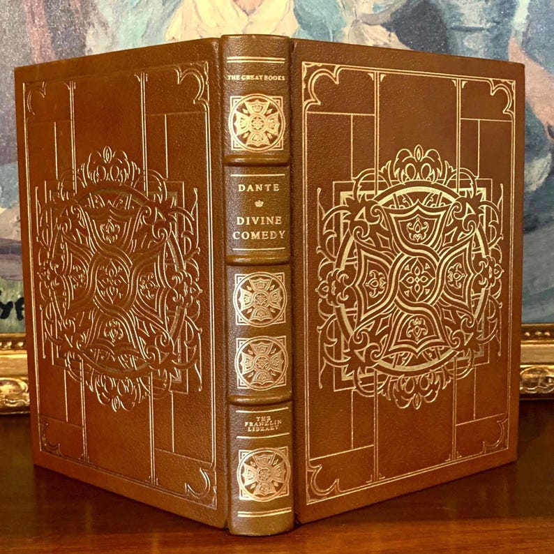 Leather-bound edition of Dante's 'Divine Comedy' from The Franklin Library, featuring an ornate gold-embossed design on the cover with intricate patterns and symbols, displayed against a backdrop of a framed painting.