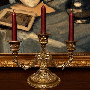 Puede incluir: Un candelabro de metal dorado con tres portavelas que presentan detalles ornamentales en volutas y una base circular ancha. Tres velas cónicas de color burdeos intenso están insertadas en los soportes, dispuestas de forma simétrica.