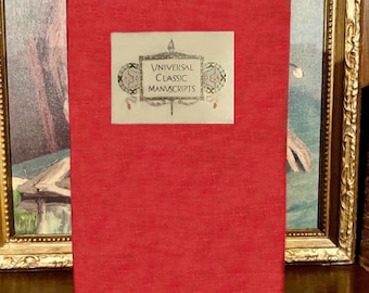 May include: A red book with the title "Universal Classic Manuscripts" in a white rectangular frame. The book has a textured cover and is standing upright. The background includes a framed painting.