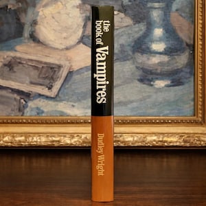 May include: A hardcover book titled "the book of Vampires" by Dudley Wright. The spine is black with white text, transitioning to a tan color. The book is standing upright against a backdrop of a framed painting.