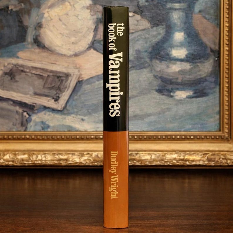 May include: A hardcover book titled "the book of Vampires" by Dudley Wright. The spine is black with white text, transitioning to a tan color. The book is standing upright against a backdrop of a framed painting.