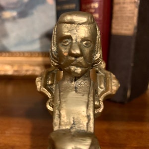 May include: A vintage brass door knocker featuring a detailed bust of a man with a mustache and sideburns. The knocker has an aged patina, suggesting it is antique. The design is ornate, with decorative elements around the bust.