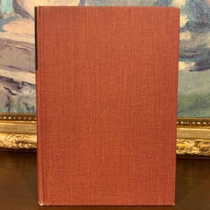 May include: A hardcover book with a textured, reddish-brown cover. The book is standing upright, with the spine facing left. The background includes a framed painting with a gold frame.