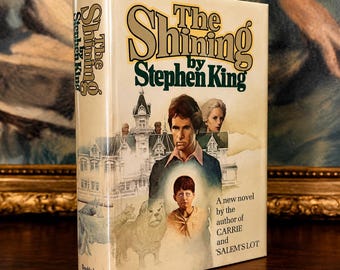 May include: A hardcover book titled The Shining by Stephen King with a cream-colored dust jacket. The cover features an illustration of a large house, a man, a woman, a child with blank eyes, and a lion. Text includes A new novel by the author of CARRIE and SALEM'S LOT.