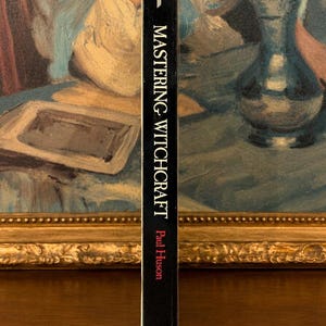Puede incluir: Un libro negro titulado "Mastering Witchcraft" de Paul Huson. El lomo del libro es visible, con texto blanco sobre fondo negro. El libro est&aacute; situado frente a una pintura con un marco dorado.