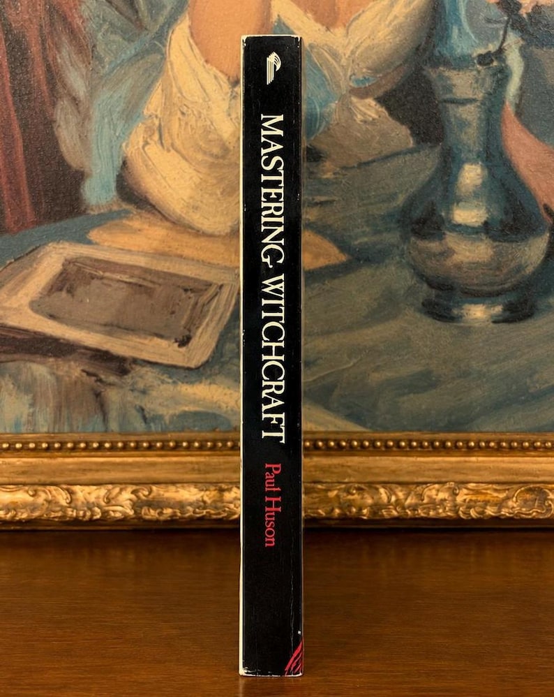 Puede incluir: Un libro negro titulado "Mastering Witchcraft" de Paul Huson. El lomo del libro es visible, con texto blanco sobre fondo negro. El libro est&aacute; situado frente a una pintura con un marco dorado.