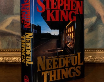 May include: A hardcover book titled Needful Things by Stephen King. The cover features a dark street scene at dusk with a storefront window displaying an Open sign. The author's name is in large red letters, and the title is in bold gold text.