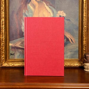 May include: A hardcover book with a textured red cover stands on a wooden surface. The book is centered in the frame, with a painting in a gold frame visible in the background. The painting depicts a woman in a blue dress.