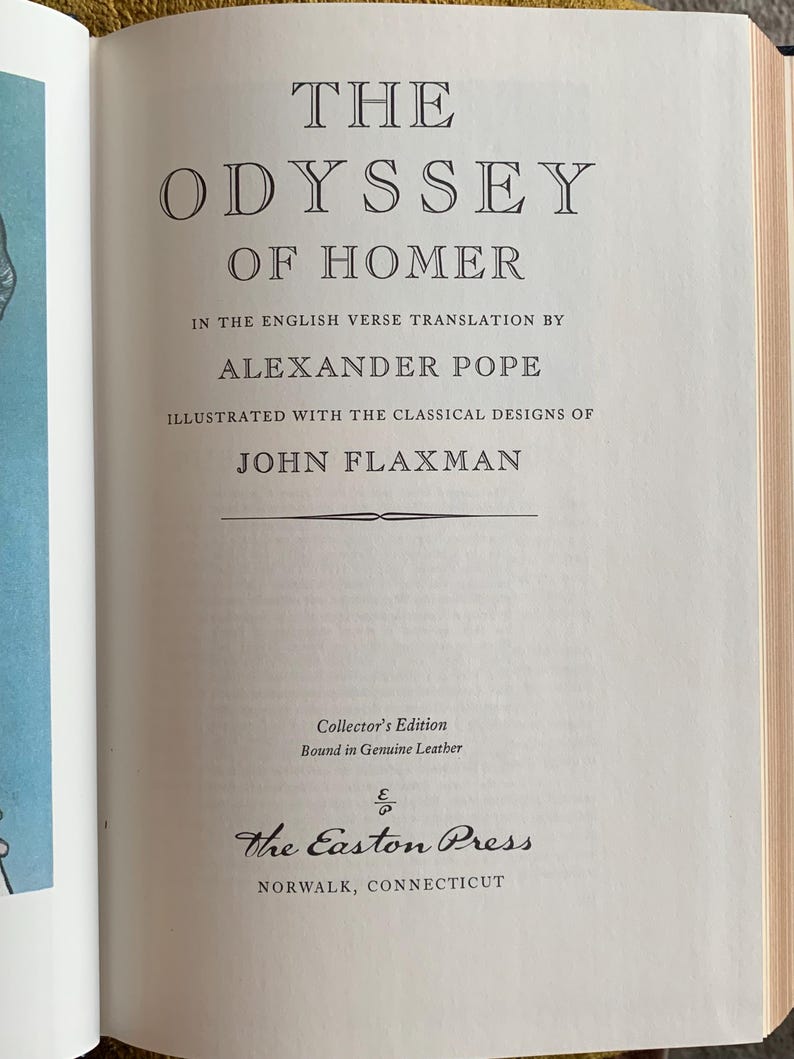 以下が含まれることがあります： 開かれた本のページ。タイトルは「The Odyssey of Homer」で、セリフ体で表示されています。翻訳者アレクサンダー・ポープ、イラストレーターのジョン・フラックスマンの名前も記載。コレクターズエディションで、本革装丁、The Easton Press発行。