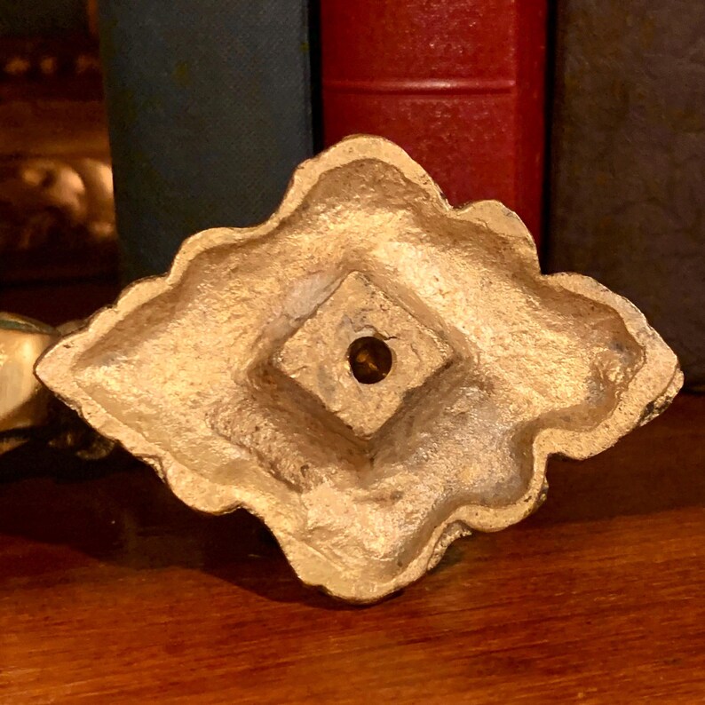 May include: A gold-colored, ornate decorative base with a diamond-shaped center and a scalloped edge. The base has a textured surface and a small hole in the center. The background includes a wooden surface and the spines of books.