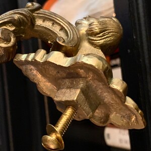 May include: Close-up of a gold-colored furniture handle with an ornate design. The handle features a sculpted face and decorative flourishes. A brass screw is attached to the handle. The handle is likely for a cabinet or drawer.
