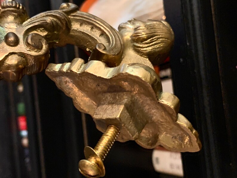 May include: Close-up of a gold-colored furniture handle with an ornate design. The handle features a sculpted face and decorative flourishes. A brass screw is attached to the handle. The handle is likely for a cabinet or drawer.