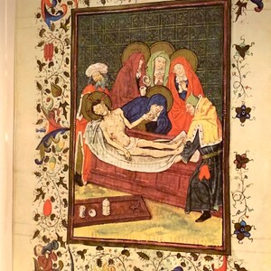 A medieval painting depicting the lamentation of Christ. The painting is framed in a gold frame with a decorative border of vines and flowers. The painting shows a group of people gathered around the body of Christ, who is lying on a wooden table. The people are dressed in colorful robes and are expressing their grief. The painting is done in a realistic style and is full of detail.