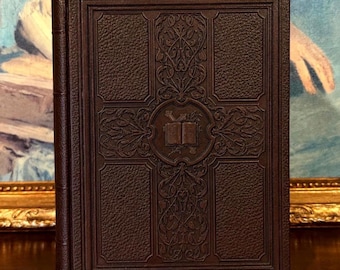 May include: A dark brown hardcover book featuring a textured leather-like finish with an embossed cross design. The center of the cover displays an intricate illustration of an open book surrounded by detailed floral and vine scrollwork patterns.
