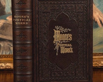May include: A dark brown leather-bound book titled Moore's Poetical Works. The cover features intricate embossed patterns and gold-stamped lettering. The spine includes raised bands with decorative gold-colored floral motifs and the title.