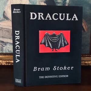 May include: A black hardcover book with the title "Dracula" in silver lettering. The author, Bram Stoker, is also printed in silver lettering. The book cover features a red background with a black silhouette of a figure in a cape.