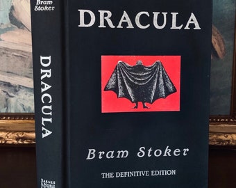 Puede incluir: Un libro negro de tapa dura con el título "Dracula" en letras plateadas. El autor, Bram Stoker, también está impreso en letras plateadas. La portada del libro presenta un fondo rojo con una silueta negra de una figura con una capa.