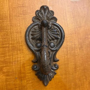 May include: Dark brown cast iron door knocker with an elaborate floral design. The knocker has a central handle and a detailed, symmetrical pattern, ideal for adding a touch of vintage charm to any door.