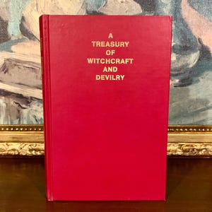 Puede incluir: Un libro de tapa dura rojo titulado "A Treasury of Witchcraft and Devilry" en letras doradas. El libro est&aacute; de pie, con el t&iacute;tulo en la portada. El fondo presenta una pintura borrosa.