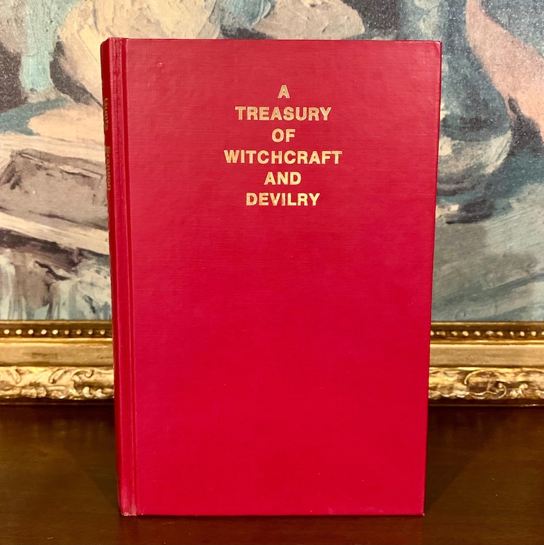 Puede incluir: Un libro de tapa dura rojo titulado "A Treasury of Witchcraft and Devilry" en letras doradas. El libro est&aacute; de pie, con el t&iacute;tulo en la portada. El fondo presenta una pintura borrosa.