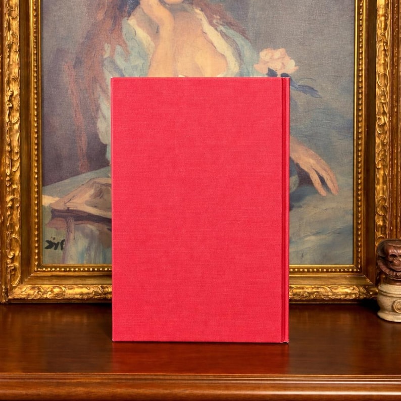 May include: A hardcover book with a bright red cover stands upright on a wooden surface. The book's spine is visible, and the cover has a textured appearance. The background features a framed painting with gold trim.