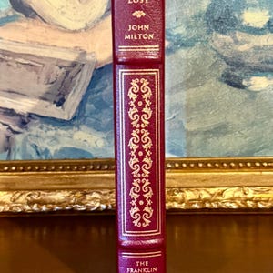 Może przedstawiać: Burgundowa książka w sk&oacute;rzanej oprawie zatytułowana "Paradise Lost" autorstwa Johna Miltona. Grzbiet zdobią złote zdobienia i napis "The Franklin Library". Książka jest wyświetlana pionowo.