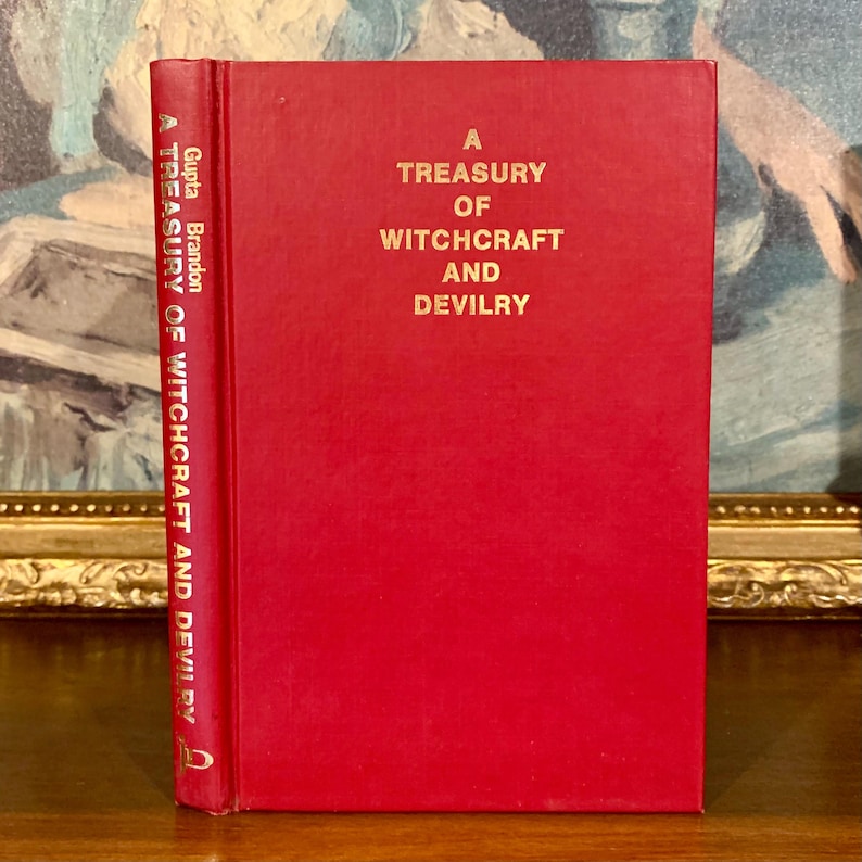 Puede incluir: Un libro rojo de tapa dura con letras doradas, titulado "A Treasury of Witchcraft and Devilry". El lomo muestra el t&iacute;tulo y el nombre del autor. El libro est&aacute; sobre una superficie de madera.