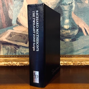 May include: A hardcover book titled "Richard Matheson's The Twilight Zone Scripts" in black with white lettering. The book has a silver logo and a signature on the side. The book is displayed in front of a painting.