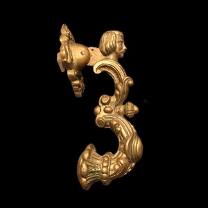 May include: Ornate brass door handle with a classical design. The handle features a profile of a man's face and intricate scrollwork. The handle is a warm gold color and is set against a black background.