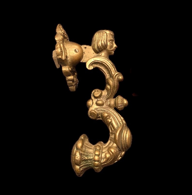 May include: Ornate brass door handle with a classical design. The handle features a profile of a man's face and intricate scrollwork. The handle is a warm gold color and is set against a black background.