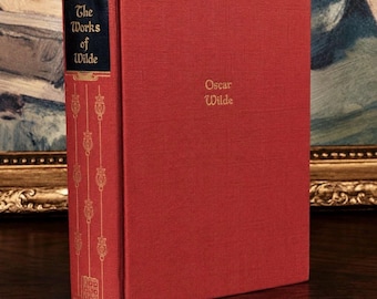May include: A red hardcover book titled The Works of Wilde by Oscar Wilde. The spine features a dark blue label with gold lettering and vertical gold decorative lines. The front cover displays the author's name in gold text centered on the red fabric.