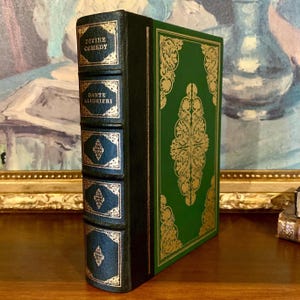 Puede incluir: Un libro de tapa dura con una cubierta verde y un diseño de filigrana dorada. El lomo es azul oscuro con letras doradas que dicen "DIVINE COMEDY" y "DANTE ALIGHIERI". El libro se exhibe sobre una superficie de madera.