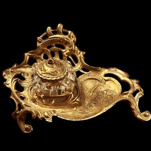May include: A gold-toned metal inkwell with a glass ink pot and a decorative base. The base is ornately designed with swirls and scrolls.