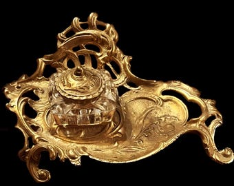 May include: A gold-toned metal inkwell stand featuring an ornate, swirling openwork frame with a clear glass ink container at the center. The glass inkwell has a matching gold-toned metal lid with a pointed finial and decorative leaf-like engravings.