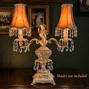 K&ouml;nnte beinhalten: Eine zweiflammige Tischleuchte mit einem cremefarbenen Metallsockel, der eine Puttenfigur mit einem Dreizack zeigt. Die Lampe hat ein Mittelteil aus geschliffenem Glas, h&auml;ngende klare Kristallprismen und zwei orangefarbene Stoffschirme mit schwarzem Drahtdekor.