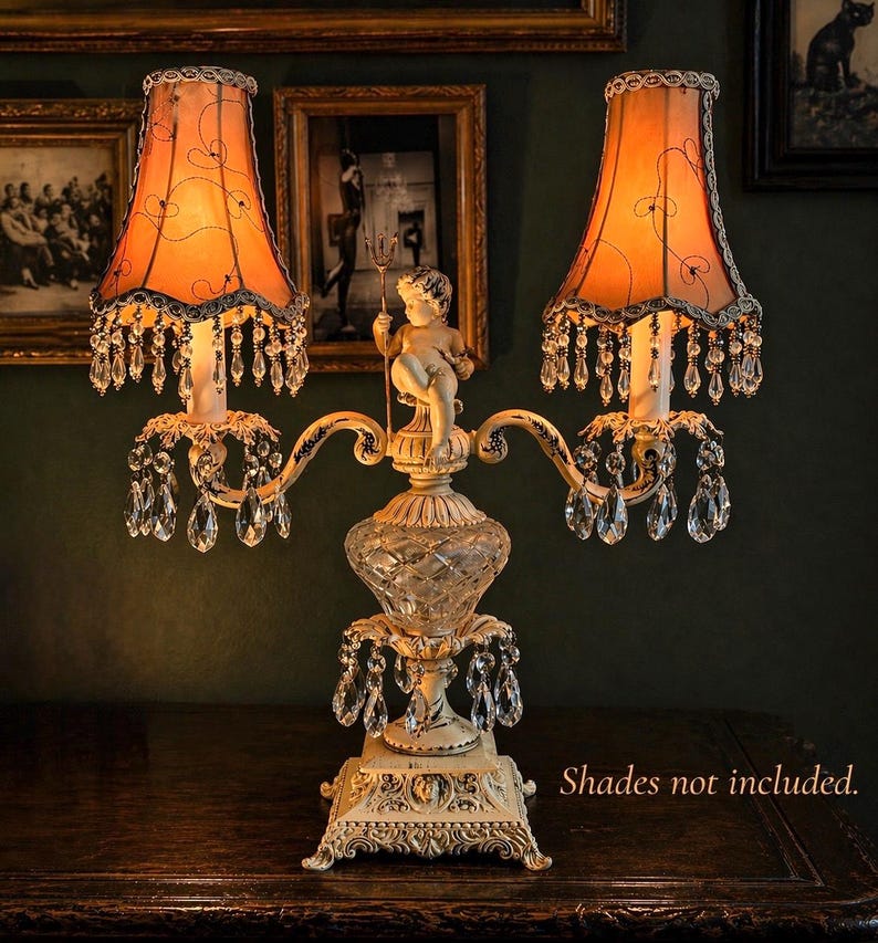 K&ouml;nnte beinhalten: Eine zweiflammige Tischleuchte mit einem cremefarbenen Metallsockel, der eine Puttenfigur mit einem Dreizack zeigt. Die Lampe hat ein Mittelteil aus geschliffenem Glas, h&auml;ngende klare Kristallprismen und zwei orangefarbene Stoffschirme mit schwarzem Drahtdekor.