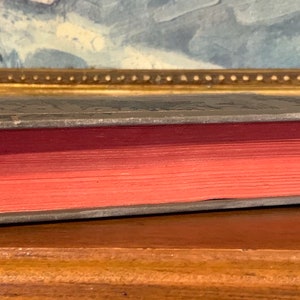 May include: Side view of an antique book with a dark gray cover and red-toned pages. The book's edges are silver, and the cover has embossed details. The book is resting on a wooden surface, with a painting in the background.