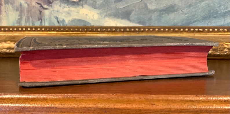 May include: Side view of an antique book with a dark gray cover and red-toned pages. The book's edges are silver, and the cover has embossed details. The book is resting on a wooden surface, with a painting in the background.